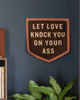 A dark brown wooden shield-shaped wall sign with beige text reading 'LET LOVE KNOCK YOU ON YOUR ASS', mounted on a dark blue wall above a green plant, with a bookshelf visible on the left.